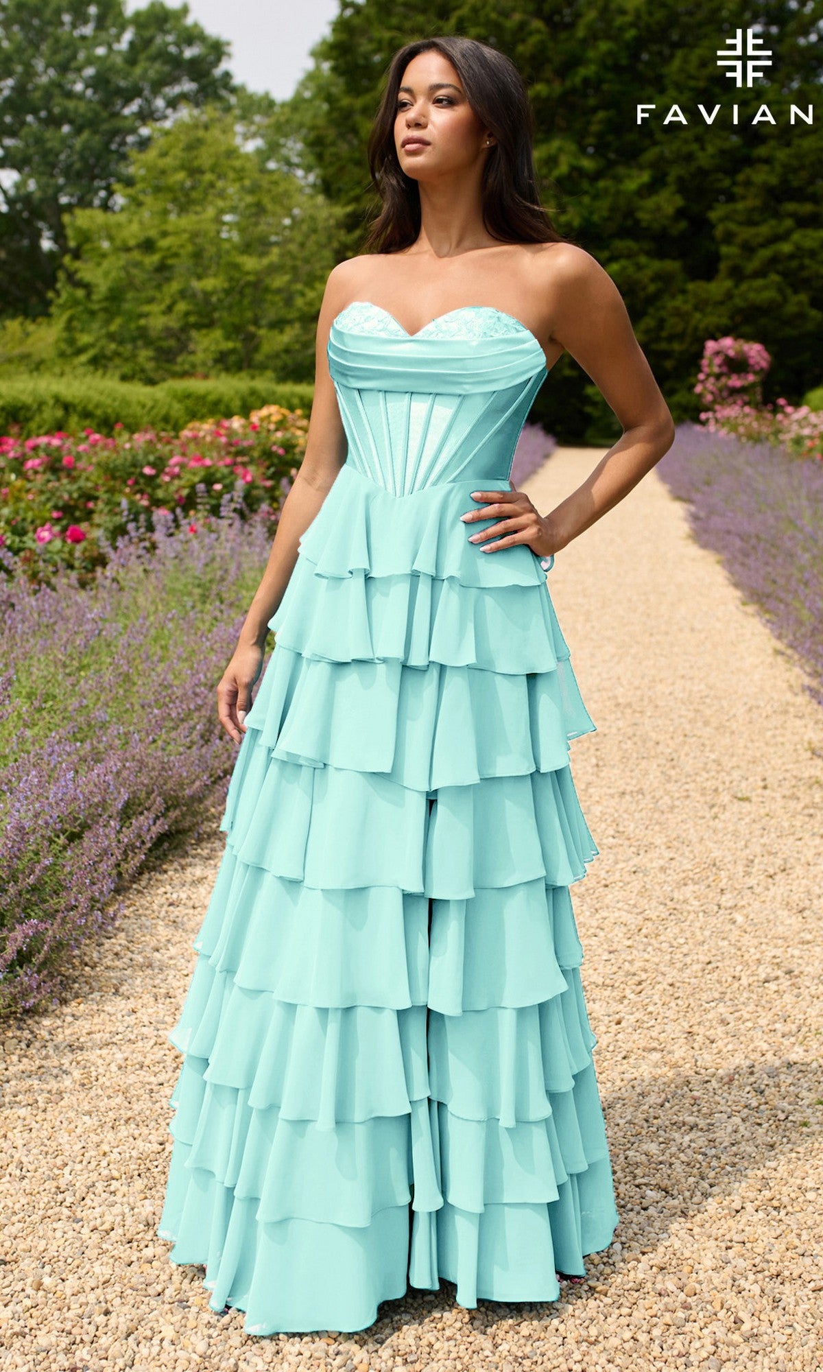 A woman stands on a flower-lined garden path wearing the Faviana 11436 Strapless Long Ruffled Prom Gown—an aqua blue, tiered dress with a sweetheart neckline, fitted bodice, and flared skirt. The FAVIAN brand name appears in the corner.